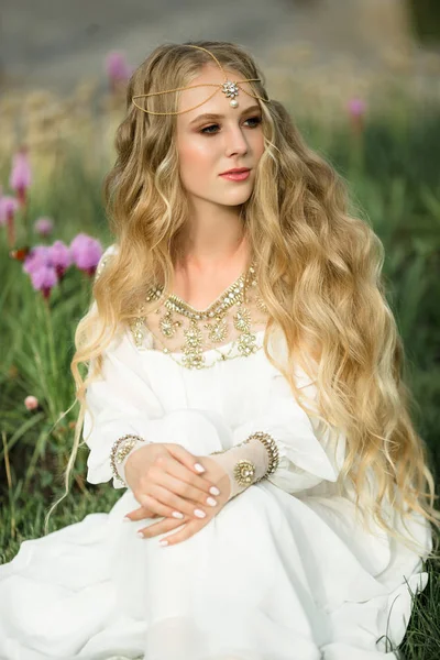 Retrato de chica rubia sonriente feliz con el pelo largo está sentado en la hierba en el parque de primavera con flores púrpuras — Foto de Stock