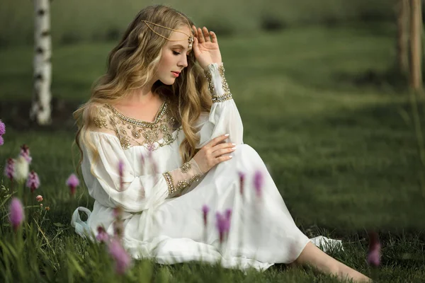 Menina loira sorridente feliz com cabelos longos está sentado na grama no parque da primavera com flores roxas — Fotografia de Stock
