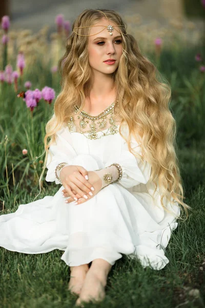 Szczęśliwy uśmiechający się blondynka z długimi włosami siedzi na trawie w parku wiosną fioletowe kwiaty — Zdjęcie stockowe