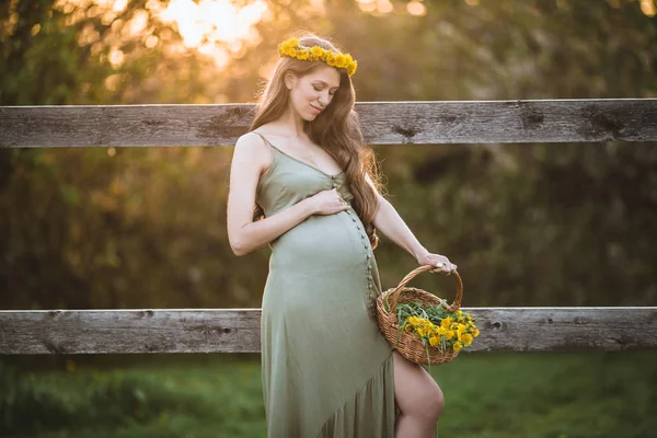 Pregnant pretty woman is wearing wreath of dandelion flowers over sunset lights , spring time — Stock Photo, Image