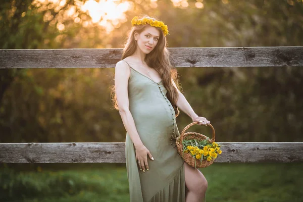 Pregnant pretty woman is wearing wreath of dandelion flowers over sunset lights , spring time — Stock Photo, Image