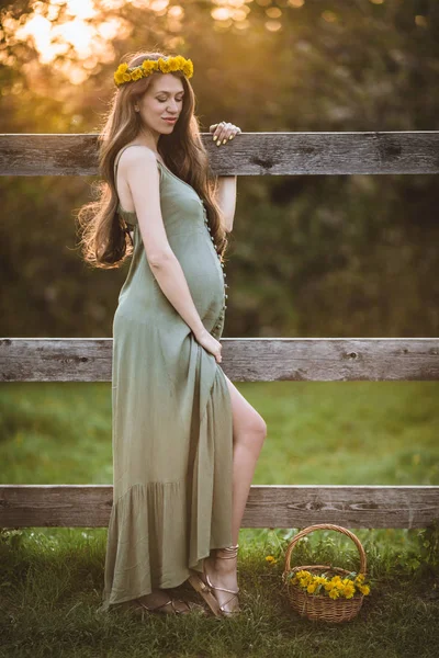 Pregnant pretty woman is wearing wreath of dandelion flowers over sunset lights , spring time — Stock Photo, Image