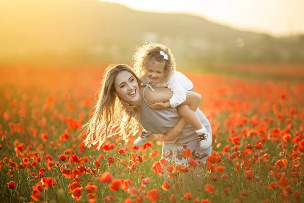 Menina bonita criança com mãe jovem estão se divertindo no campo de flores de papoula sobre luzes do pôr do sol — Fotografia de Stock