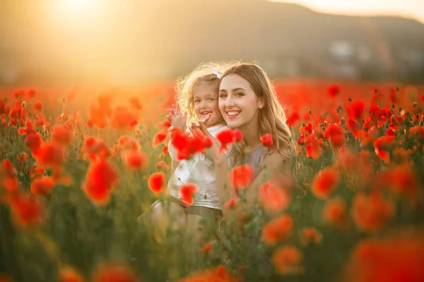 Bela menina sorridente com mãe jovem estão se divertindo no campo de flores de papoula sobre luzes do pôr do sol — Fotografia de Stock
