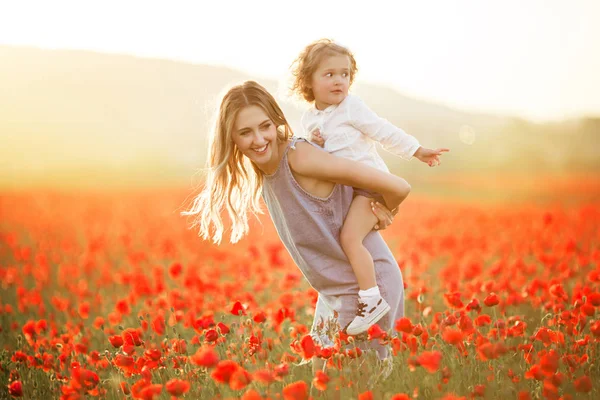Bela menina sorridente com a mãe estão se divertindo no campo de flores de papoula vermelha sobre as luzes do pôr do sol — Fotografia de Stock