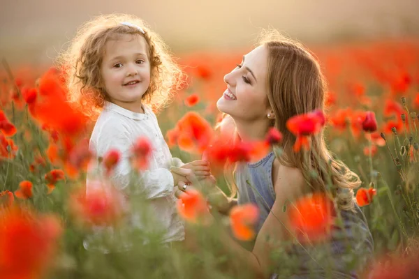 Hermosa niña sonriente con madre se divierten en el campo de las flores de amapola roja sobre las luces del atardecer —  Fotos de Stock