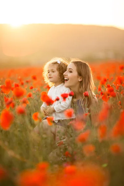Bela menina sorridente com a mãe estão se divertindo no campo de flores de papoula vermelha sobre luzes de pôr do sol, tempo de primavera — Fotografia de Stock
