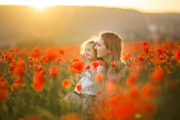 Bela menina sorridente com a mãe estão se divertindo no campo de flores vermelhas da papoula sobre as luzes do pôr do sol, tempo da primavera — Fotografia de Stock