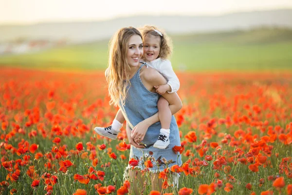 Bela menina sorridente com a mãe estão se divertindo no campo de flores vermelhas da papoula sobre as luzes do pôr do sol, tempo da primavera — Fotografia de Stock