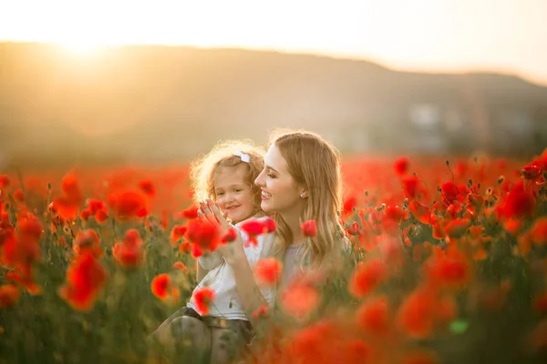 Bela menina sorridente com a mãe estão se divertindo no campo de flores vermelhas da papoula sobre as luzes do pôr do sol, tempo da primavera — Fotografia de Stock