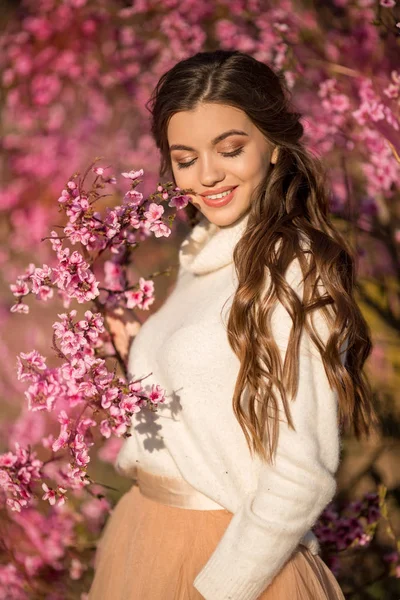 Hübsches lächelndes Mädchen posiert in der Nähe von Blütenbaum mit rosa Blumen. — Stockfoto