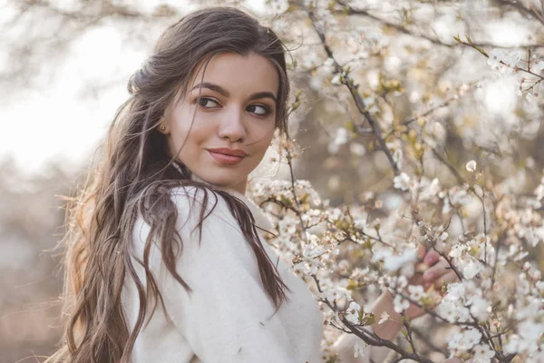 Hübsche lächelnde Teenie-Mädchen posiert in der Nähe blühen Kirschbaum mit weißen Blumen. — Stockfoto