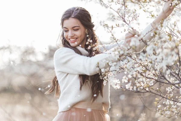 Hübsche lächelnde Teenie-Mädchen posieren im Garten in der Nähe blühender Kirschbäume mit weißen Blüten. Frühlingszeit — Stockfoto