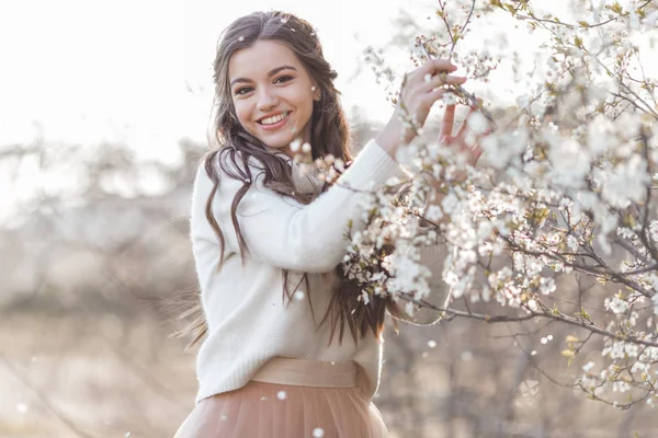 Hübsche lächelnde Teenie-Mädchen posieren im Garten in der Nähe blühender Kirschbäume mit weißen Blüten. Frühlingszeit — Stockfoto