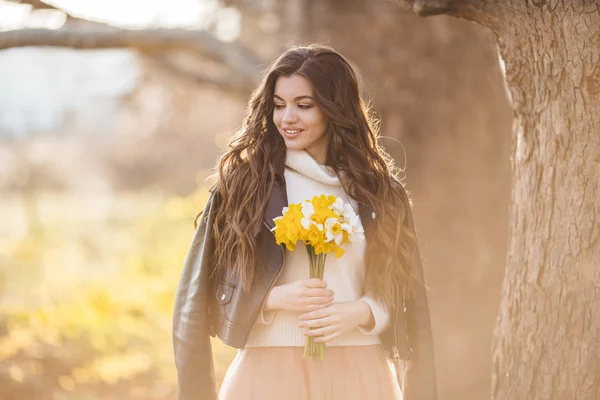 Pretty smiling teen girl are holding bouquet of narcissus flowers over sunset lights. Spring time