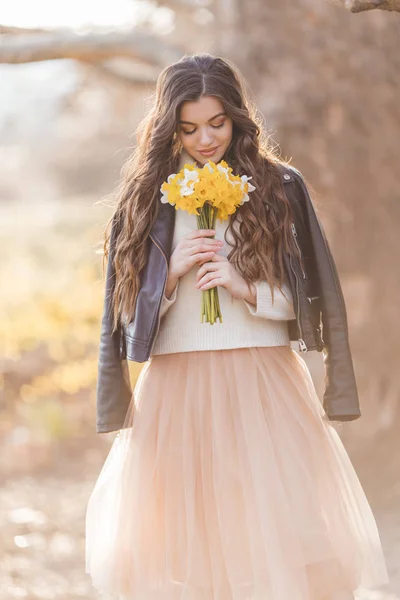 Pretty smiling teenager girl are holding bouquet of narcissus flowers in park over sunset lights. Spring time