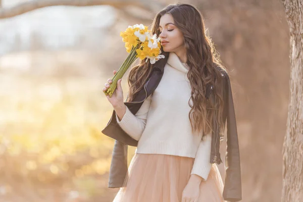 Pretty smiling teenager girl are holding bouquet of narcissus flowers in park over sunset lights. Spring time