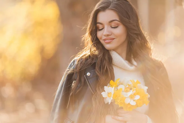 Pretty smiling teenager girl are holding bouquet of narcissus flowers in park over sunset lights. Spring time