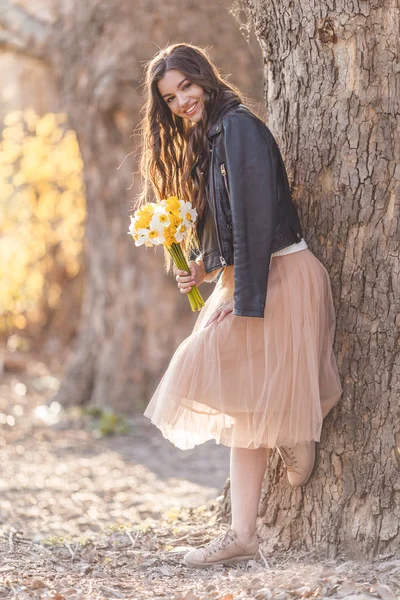 Portrait of smiling teenager girl are wearing casual clothes holding bouquet of narcissus flowers in park over sunset lights. Spring time