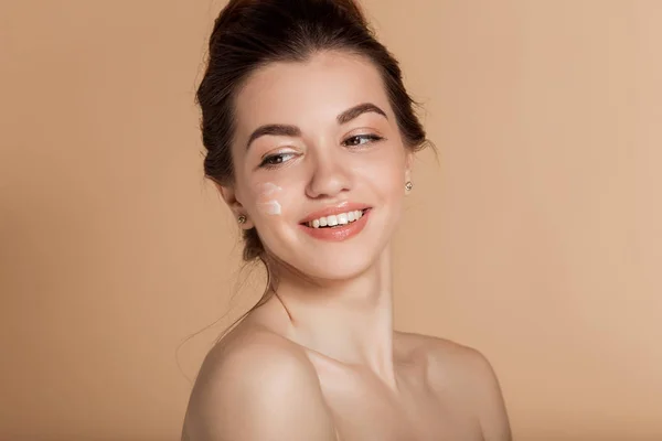 Hermoso retrato facial de una joven sonriente con crema cosmética en la mejilla. Cuidado de la piel y concepto de salud . — Foto de Stock