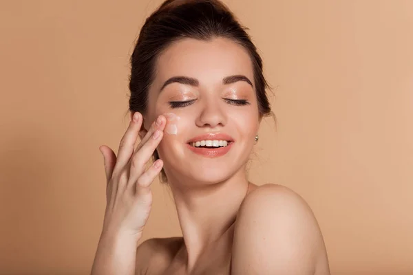 Beautiful face portrait of young woman is applying face cream on a cheek. Skin care and health concept. — Stock Photo, Image