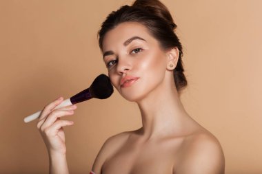 Closeup portrait of pretty girl with makeup brush in hand. Isolated on beige. Beauty and cosmetics concept.