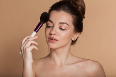 Closeup portrait of beautiful young girl with perfect skin is holding makeup brush in hand. Isolated on beige. Beauty and cosmetics concept.