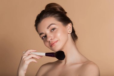 Closeup portrait of beautiful happy young girl with perfect skin is holding makeup brush in hand. Isolated on beige. Beauty and cosmetics concept.
