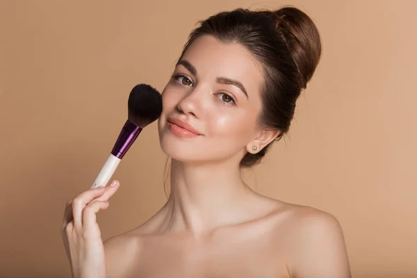 Primer plano retrato de hermosa joven feliz con la piel perfecta está sosteniendo cepillo de maquillaje en la mano. Aislado en beige. Concepto de belleza y cosmética . —  Fotos de Stock