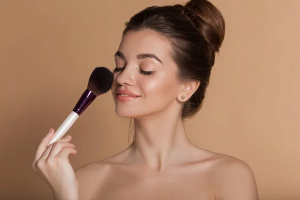 Closeup portrait of beautiful happy young girl with perfect skin is holding makeup brush in hand. Isolated on beige. Beauty and cosmetics concept. — Stock Photo, Image