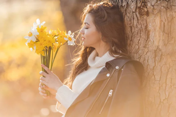 笑顔のティーンエイジャーの女の子の肖像画は、日没の光の上に公園でナルキッソスの花の花束を保持しています。春の時間 — ストック写真