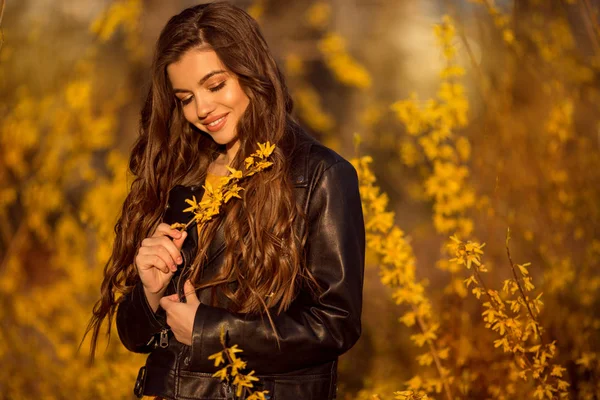 Porträt eines lächelnden Teenager-Mädchens mit langen Haaren steht in der Nähe von Büschen mit gelben Blumen. Frühlingszeit. — Stockfoto