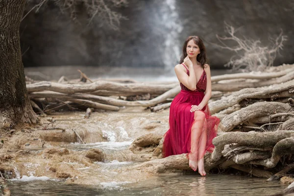 Fiatal gyönyörű nő visel divat piros ruha ül az erdőben, közel a vízesés — Stock Fotó