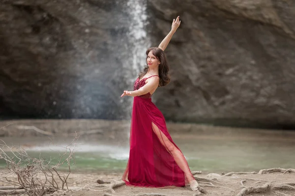 Krásná žena má na sobě módní červené šaty tančící v lese poblíž vodopádu, sport a módní koncepce. — Stock fotografie