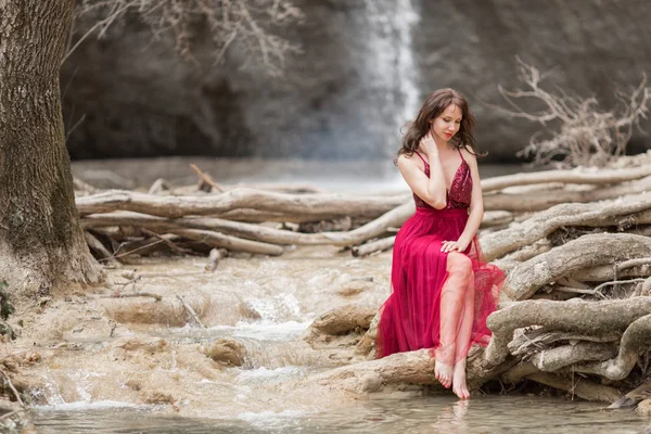 Joven hermosa mujer está usando vestido rojo de moda sentado en el bosque cerca de la cascada — Foto de Stock