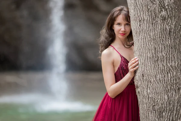 Giovane bella donna indossa abito rosso moda a piedi nella foresta vicino alla cascata — Foto Stock