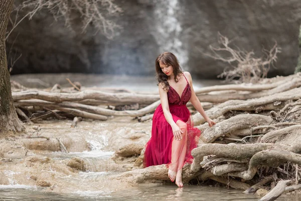 Fiatal gyönyörű nő visel divat piros ruha ül az erdőben, közel a vízesés — Stock Fotó