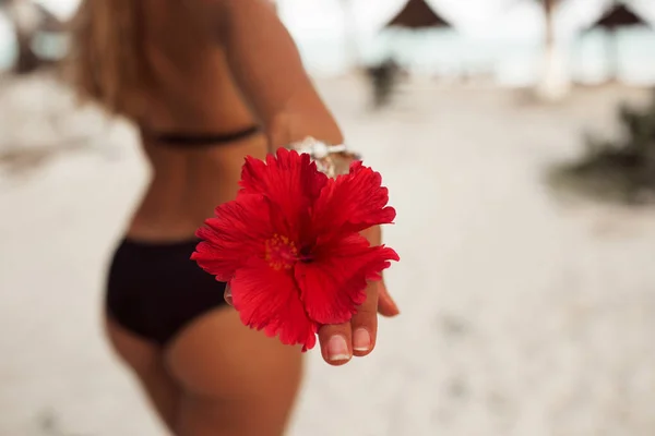 年轻的晒黑的女人与完美的臀部与纹身在她的手和红花是走在海滩上。美容与时尚理念. — 图库照片
