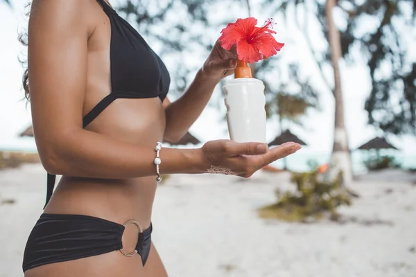 Загорелая молодая женщина с идеальным телом с татуировкой на руке и красным цветком держит банку с солнцезащитным кремом на пляже. Концепция красоты и моды . — стоковое фото