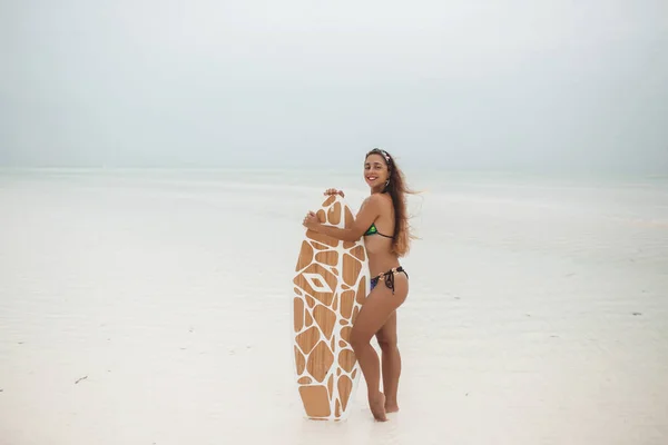 Νεαρή σέξι γυναίκα με αθλητικό μαυρισμένο σώμα, χαμογελά και τζόκινγκ με σανίδα σερφ στην παραλία σε μια όμορφη καλοκαιρινή μέρα κατά τη διάρκεια της χαμηλής παλίρροιας στη Ζανζιβάρη — Φωτογραφία Αρχείου