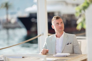 Yakışıklı işadamı deniz arka plan ile kafede oturan beyaz moda kostüm giyiyor. Karadağ körfezi.