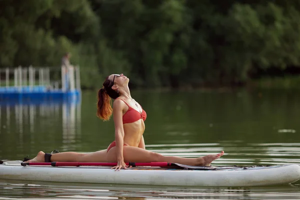 Jovem menina fitness bonita em roupa de banho está fazendo ioga no SUP no meio do lago com uma floresta verde no fundo . — Fotografia de Stock