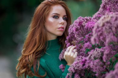 Kırmızı kıvırcık saçlı Sunshine genç gülümseyen kadın çiçeklerin mor çalı yakınında sonbahar parkı yeşil kazak ve şapka giyiyor.