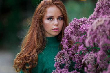 Kırmızı kıvırcık saçlı Sunshine genç gülümseyen kadın çiçeklerin mor çalı yakınında sonbahar parkı yeşil kazak ve şapka giyiyor.