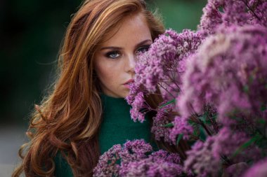 Kırmızı kıvırcık saçlı Sunshine genç gülümseyen kadın çiçeklerin mor çalı yakınında sonbahar parkı yeşil kazak ve şapka giyiyor.