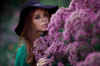 Kırmızı kıvırcık saçlı Sunshine genç gülümseyen kadın sonbahar parkta yeşil kazak ve şapka giyiyor.