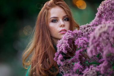 Kırmızı kıvırcık saçlı Sunshine genç gülümseyen kadın çiçeklerin mor çalı yakınında sonbahar parkı yeşil kazak ve şapka giyiyor.