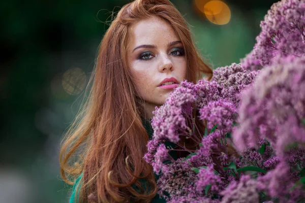 Sunshine ung leende kvinna med rött lockigt hår bär grön tröja och hatt i höst Park nära lila buske av blommor. — Stockfoto