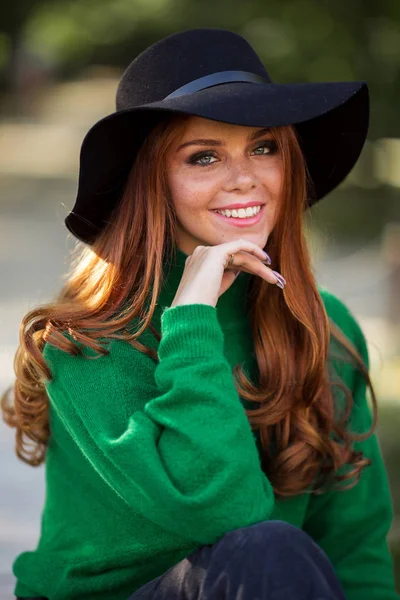 Sunshine joven sonriente mujer con el pelo rizado rojo está usando suéter verde y sombrero en el parque de otoño . — Foto de Stock