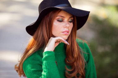 Kırmızı kıvırcık saçlı Sunshine genç genç kız sonbahar parkta yeşil kazak ve şapka giyiyor.